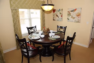 A dining room with a round dark wooden table set for four people. The table has white plates, bowls, and cups, with a decorative centerpiece featuring a ceramic chicken. Four dark wooden chairs with red cushions surround the table. The room has beige walls, two framed bird-themed artworks, a window with green patterned curtains, and a hanging light fixture above the table.