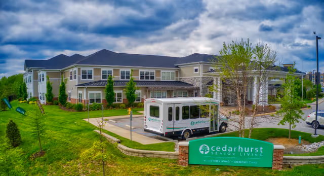 Exterior view of Cedarhurst of Des Peres senior living facility showing a two-story building with multiple windows, a driveway with a Cedarhurst shuttle bus parked, green landscaped lawns, young trees, and a green sign with the Cedarhurst logo and text. The sky is cloudy.
