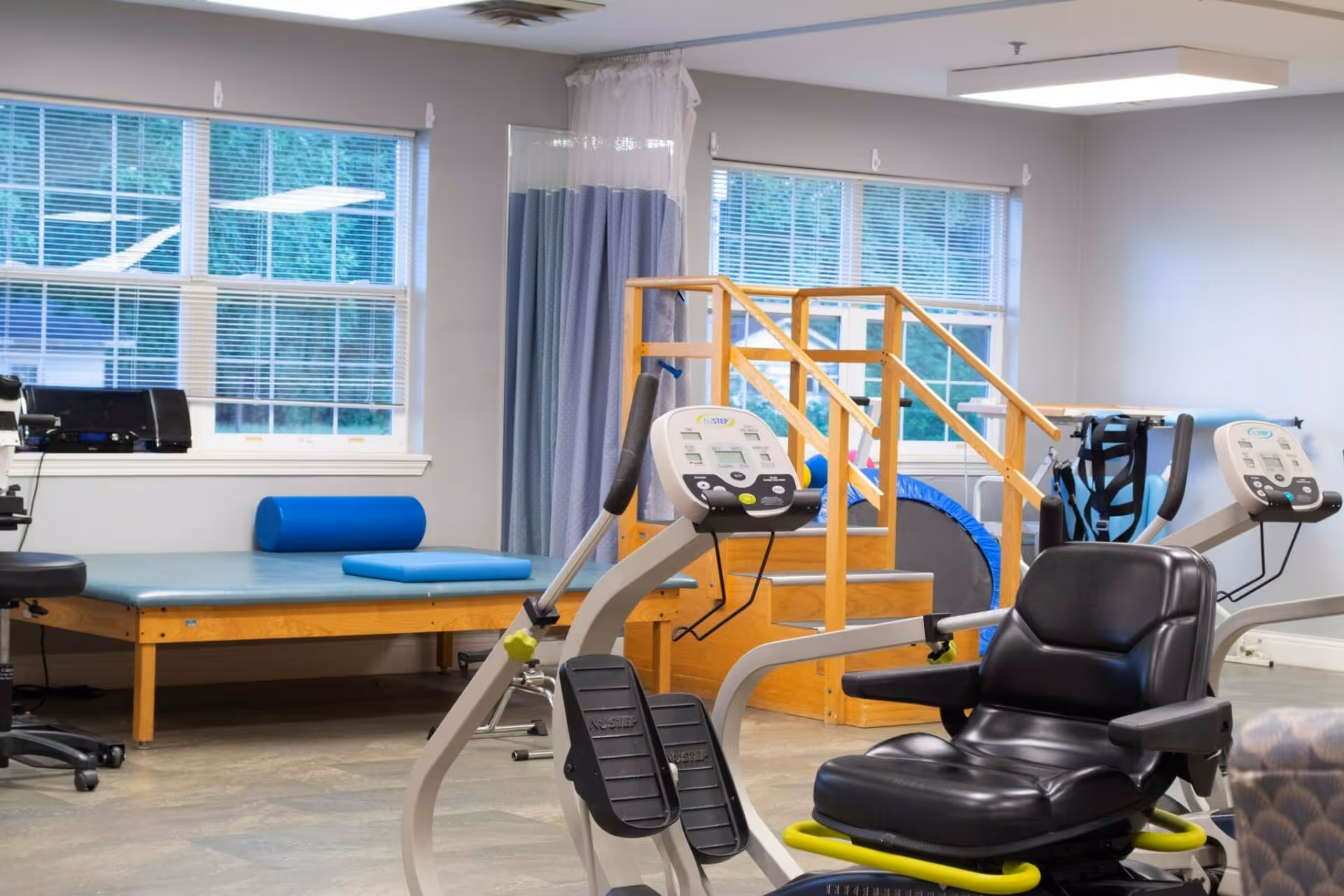A rehabilitation exercise room with a recumbent bike, therapy table, and wooden steps with handrails near windows.