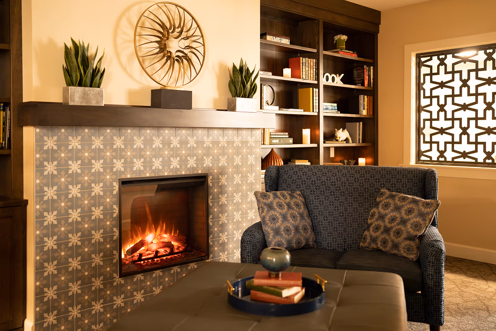 Cozy living room area with a lit fireplace surrounded by patterned tiles, a dark wooden mantel with decorative plants and a metal wall art piece above it. To the right, there is a dark blue patterned loveseat with two decorative pillows, a window with a geometric patterned screen, and a built-in wooden bookshelf filled with books and decorative items. A dark ottoman with a tray holding books and a small vase is in the foreground.