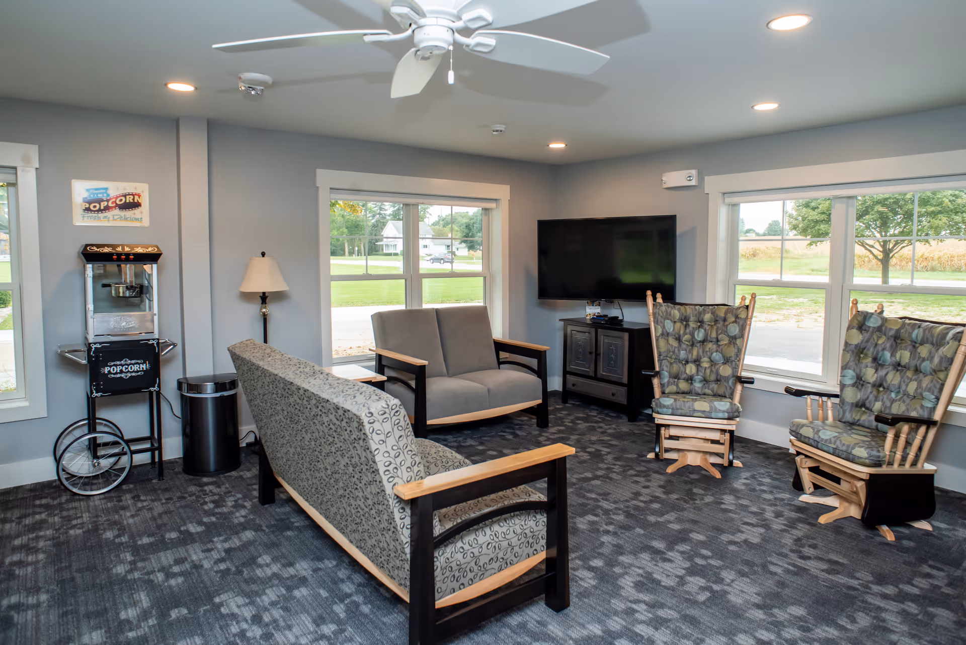 Communal sitting area with sofas and patterned armchairs facing a wall-mounted TV, large windows, and a popcorn machine.