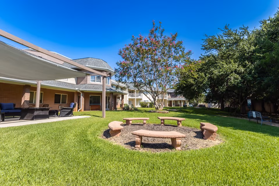 Outdoor grassy area at Asher Point Independent Living of Arlington with a circular arrangement of stone benches around a fire pit, surrounded by trees and a building with a covered patio and outdoor seating under a clear blue sky.