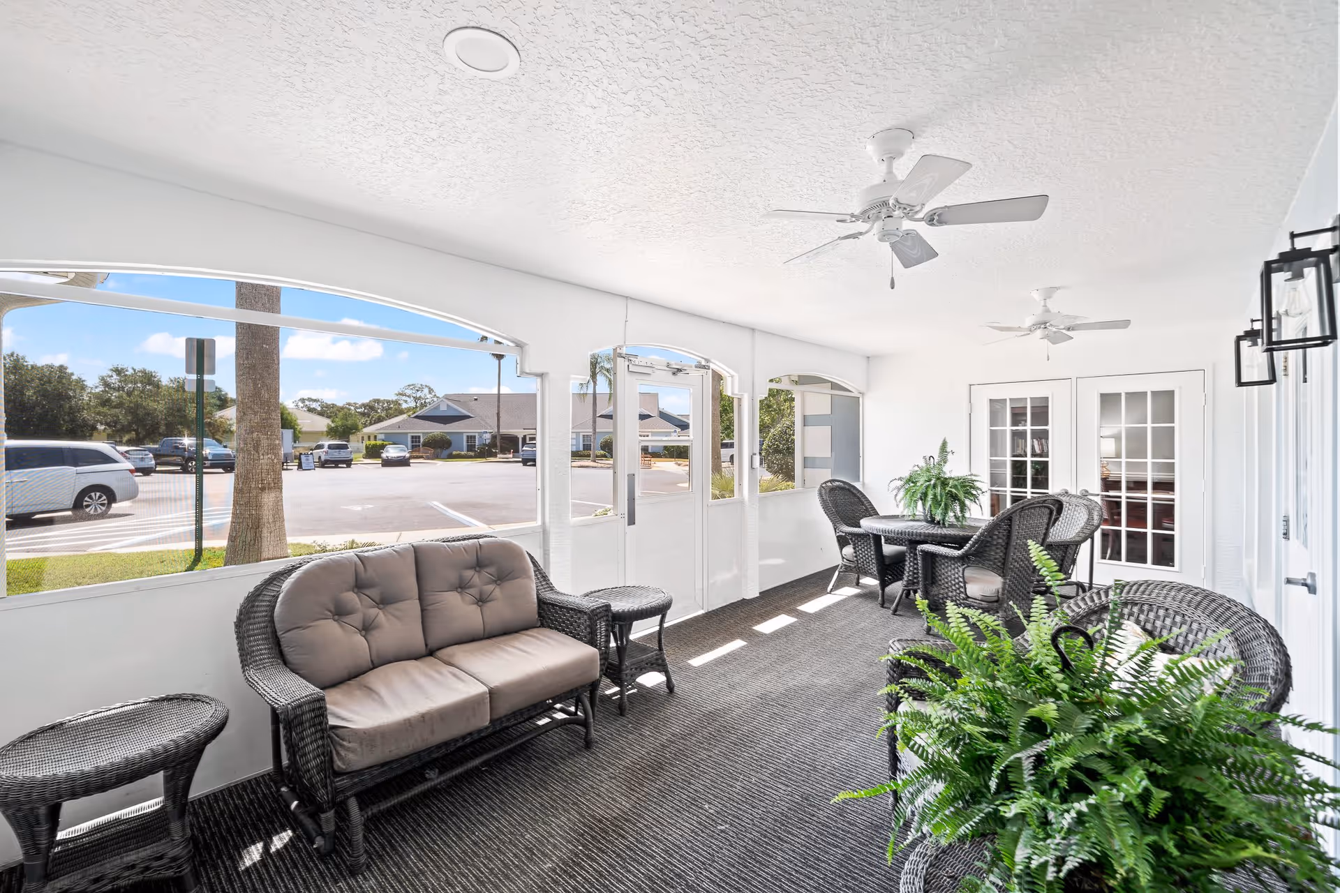 A bright and airy enclosed porch area with white walls and ceiling, two ceiling fans, and large screened windows looking out to a parking lot. The porch is furnished with dark wicker furniture including a cushioned loveseat, chairs, small tables, and a round table with chairs. There are green potted plants on the tables and floor, and double French doors lead inside the building.