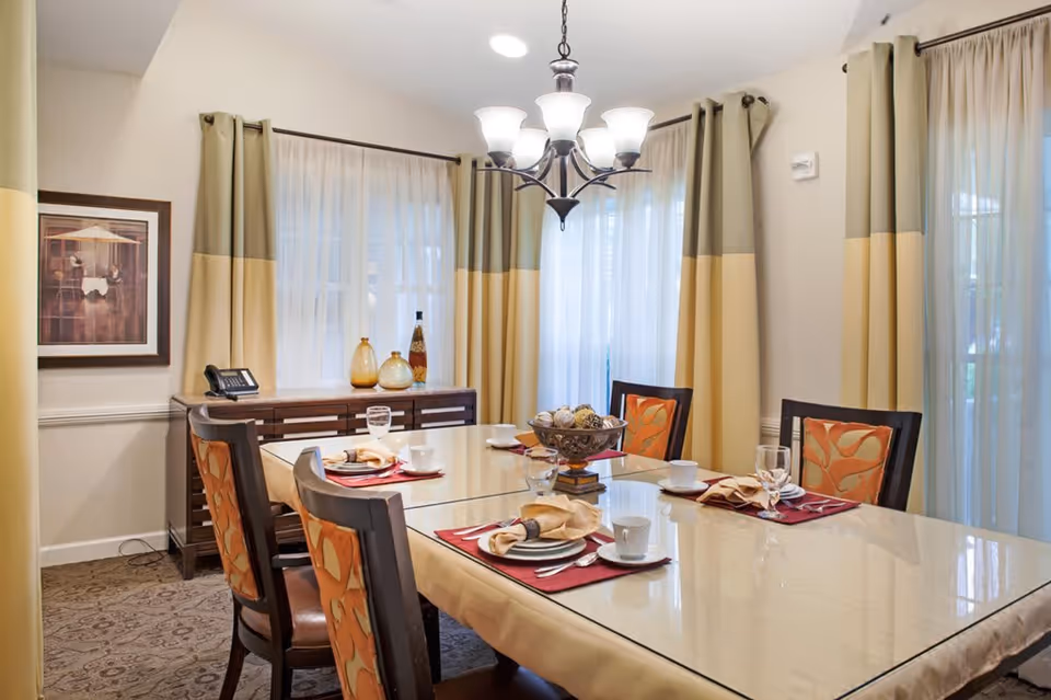 A well-lit dining room with a rectangular glass-top table set for four people. The table has beige napkins, white cups, glasses, and plates on red placemats. Four chairs with orange patterned cushions surround the table. Behind the table is a wooden sideboard with decorative vases and a telephone. The room has large windows with beige and green curtains and a chandelier hanging above the table.