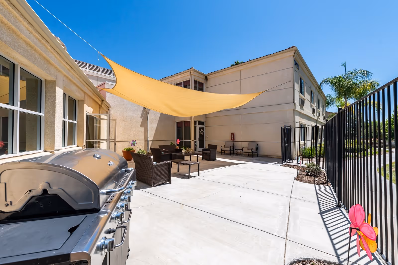 Sunlit outdoor patio with a stainless steel grill, wicker seating under a yellow shade sail, and a gated walkway.
