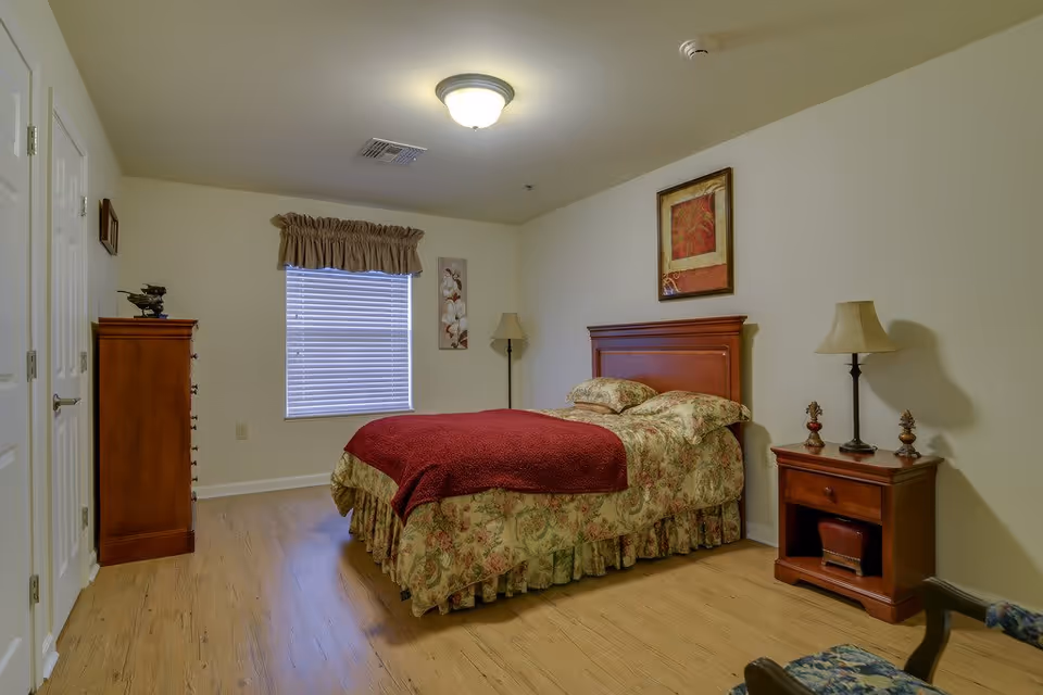 A bedroom with a wooden bed frame, floral bedspread, and a red blanket. There is a wooden nightstand with a lamp and decorative items, a wooden dresser, a window with blinds and a valance, a floor lamp, and framed artwork on the walls. The floor is light wood, and the walls are painted light beige.