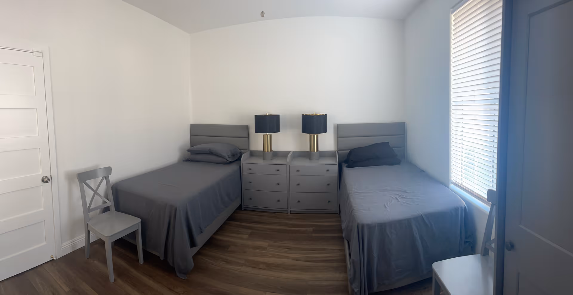 A simple bedroom with two single beds covered in gray bedding, each with a pillow. Between the beds is a gray dresser with two matching lamps featuring black shades and gold bases. There are two gray chairs, one near the door and one near the window with white blinds. The room has white walls and wooden flooring.