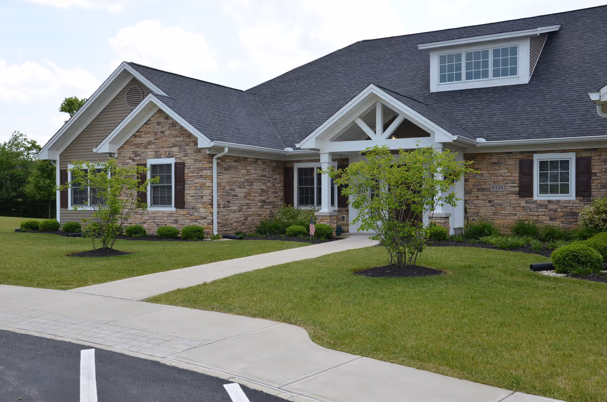 Single-story stone-faced senior living building with a covered entrance, landscaped lawn, and a paved walkway leading to the front door.