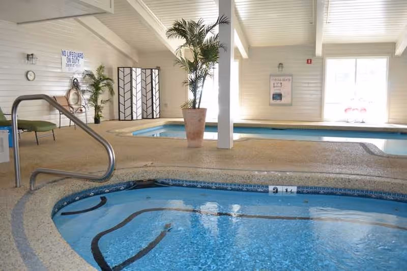Indoor pool area with a hot tub in the foreground and a swimming pool in the background. The space has beige textured flooring, white paneled walls, large windows letting in natural light, potted plants, a green chair, and a sign that reads 'No Lifeguard On Duty'.
