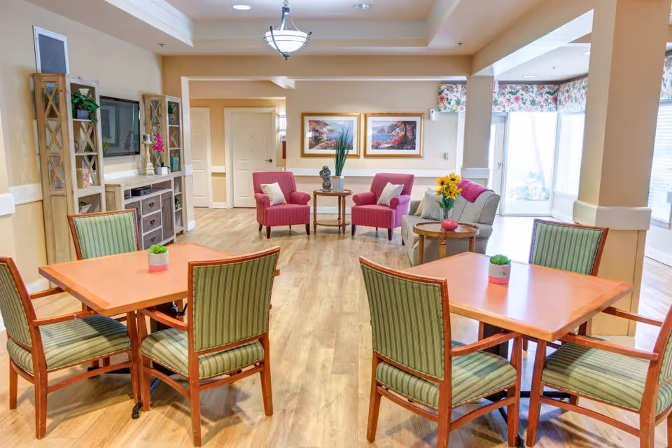 A bright and inviting common area in a senior living facility featuring two wooden tables each surrounded by four green-striped cushioned chairs. In the background, there are two pink armchairs with white pillows, a small round table with a plant, and a beige sofa with a pink throw and a vase of yellow flowers on a side table. The room has light wood flooring, beige walls, framed artwork, and large windows with floral valances letting in natural light.