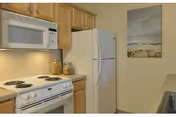 A small kitchen area with a white electric stove and oven, a white microwave mounted above the stove, wooden cabinets, a white refrigerator, and a wall painting depicting a beach scene.