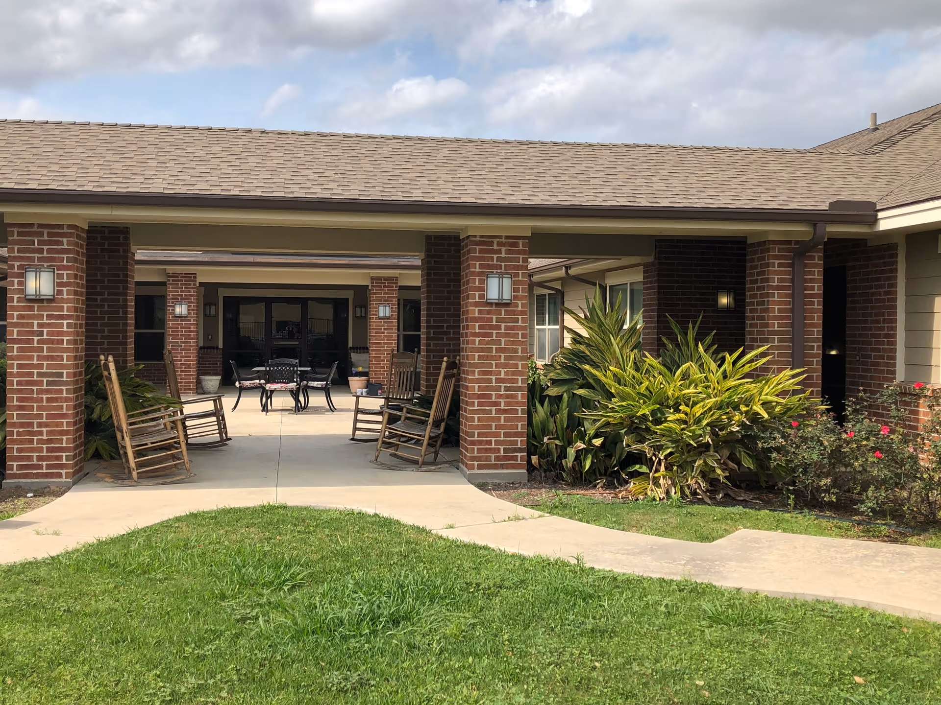 Outdoor covered patio area at a senior living facility with brick pillars, wooden rocking chairs, a table with chairs, and surrounding greenery including bushes and grass under a partly cloudy sky.