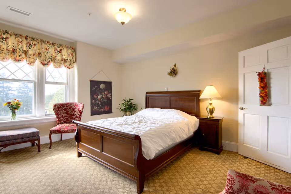 A bright bedroom with a large wooden bed covered with white bedding, a wooden nightstand with a lamp, a floral-patterned red armchair, a bench near the window, floral curtains, and a door decorated with a hanging autumn leaf ornament.