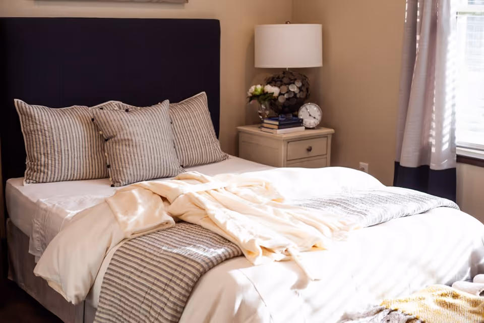 A neatly made bed with a dark upholstered headboard, three striped pillows, a beige blanket, and a cream-colored robe laid on top. Next to the bed is a small nightstand with a decorative lamp, a small clock, a vase with flowers, and a few stacked books. Light filters in through a window with curtains on the right side of the room.