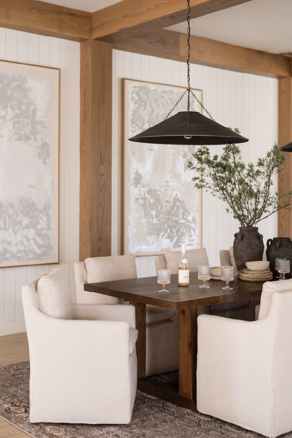 A dining area with a dark wooden table surrounded by six cushioned beige armchairs. On the table are four glasses, a bottle of wine, stacked plates, and a large vase with green branches. The room features wooden beams, white paneled walls, two abstract paintings, and a large black pendant light hanging above the table.