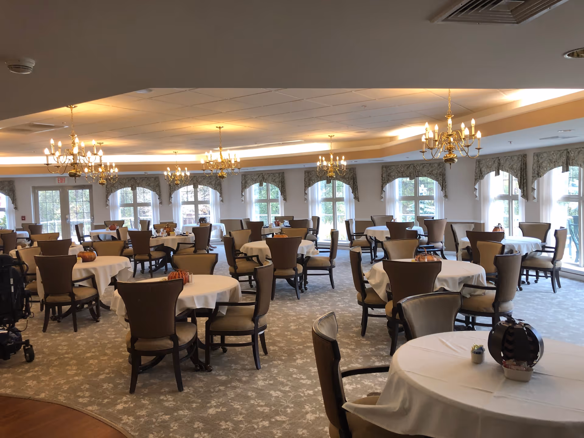 Bright, elegant dining room with round tables draped in white linens, upholstered chairs, chandeliers, and large windows.