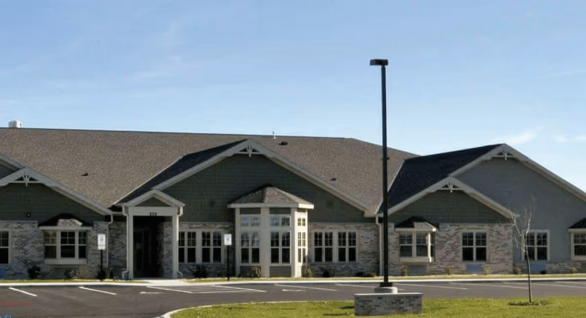 Exterior view of a single-story senior living facility building with a pitched roof, multiple windows, and a central entrance. The building is surrounded by a parking lot with marked spaces and a streetlamp in the foreground under a clear blue sky.