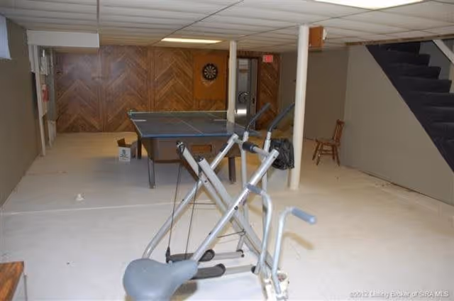 A basement recreational room with exercise equipment in the foreground, a ping pong table in the middle, a dartboard on the far wall, and a staircase on the right side. The room has a low ceiling with white tiles and wood paneling on the far wall.