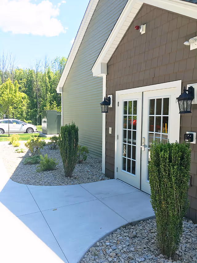 Exterior view of Beaver Lake Lodge Assisted Living showing a building entrance with double glass doors framed in white. The building has brown siding with white trim and two black wall-mounted lantern lights on either side of the doors. There is a concrete walkway leading to the entrance, bordered by landscaped areas with small bushes and rocks. In the background, there are trees and a parked white car.
