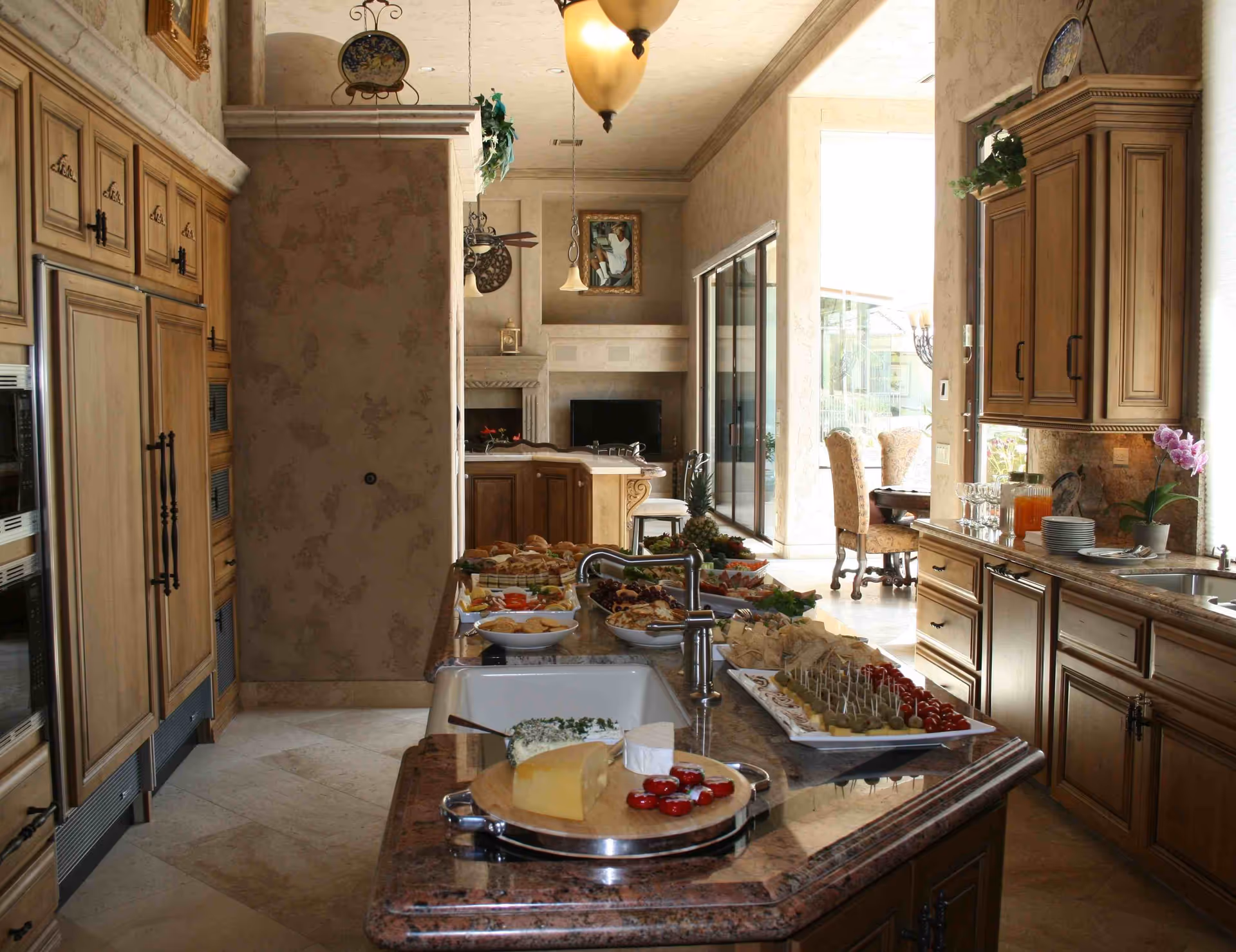 A spacious kitchen with wooden cabinets and a large granite island countertop. The countertop is set with various appetizers, cheeses, and snacks. In the background, there is a dining area with upholstered chairs and large sliding glass doors letting in natural light.