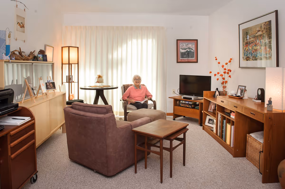 A cozy living room with a smiling elderly woman sitting in a chair near a round table with a lamp behind her. The room features a brown armchair, a wooden side table, a TV on a wooden stand, and a wooden cabinet with books and framed photos. The walls are decorated with framed artwork, and the room is softly lit with natural light coming through sheer curtains.