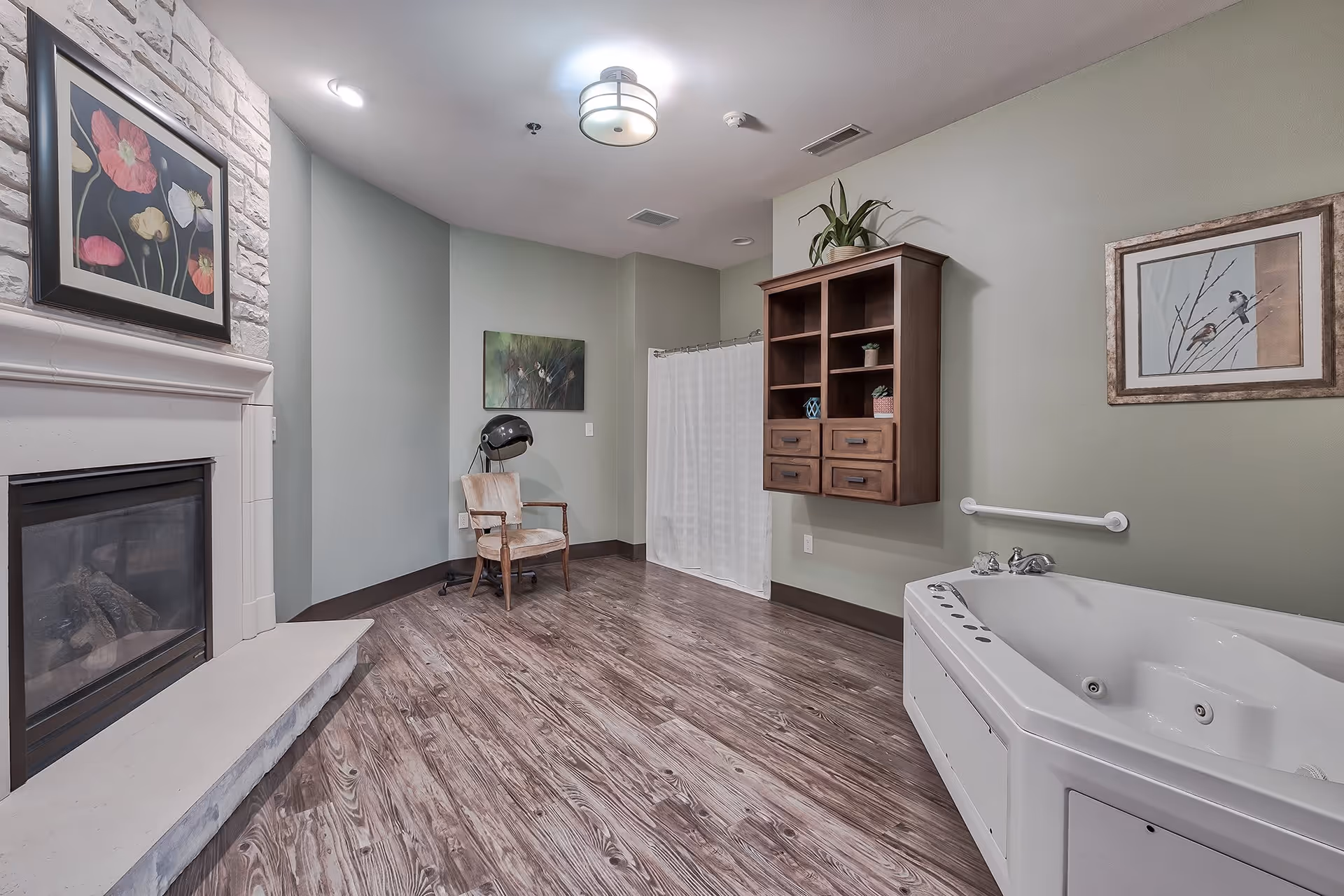 A spacious room with a large white jacuzzi tub on the right, a wooden wall cabinet above it, and a white grab bar on the wall. There is a fireplace with a white mantel and a floral painting above it on the left side. A wooden chair with a hair dryer hood is positioned near the back wall, which is painted light green. The floor is wood-patterned, and there are two framed artworks on the walls.