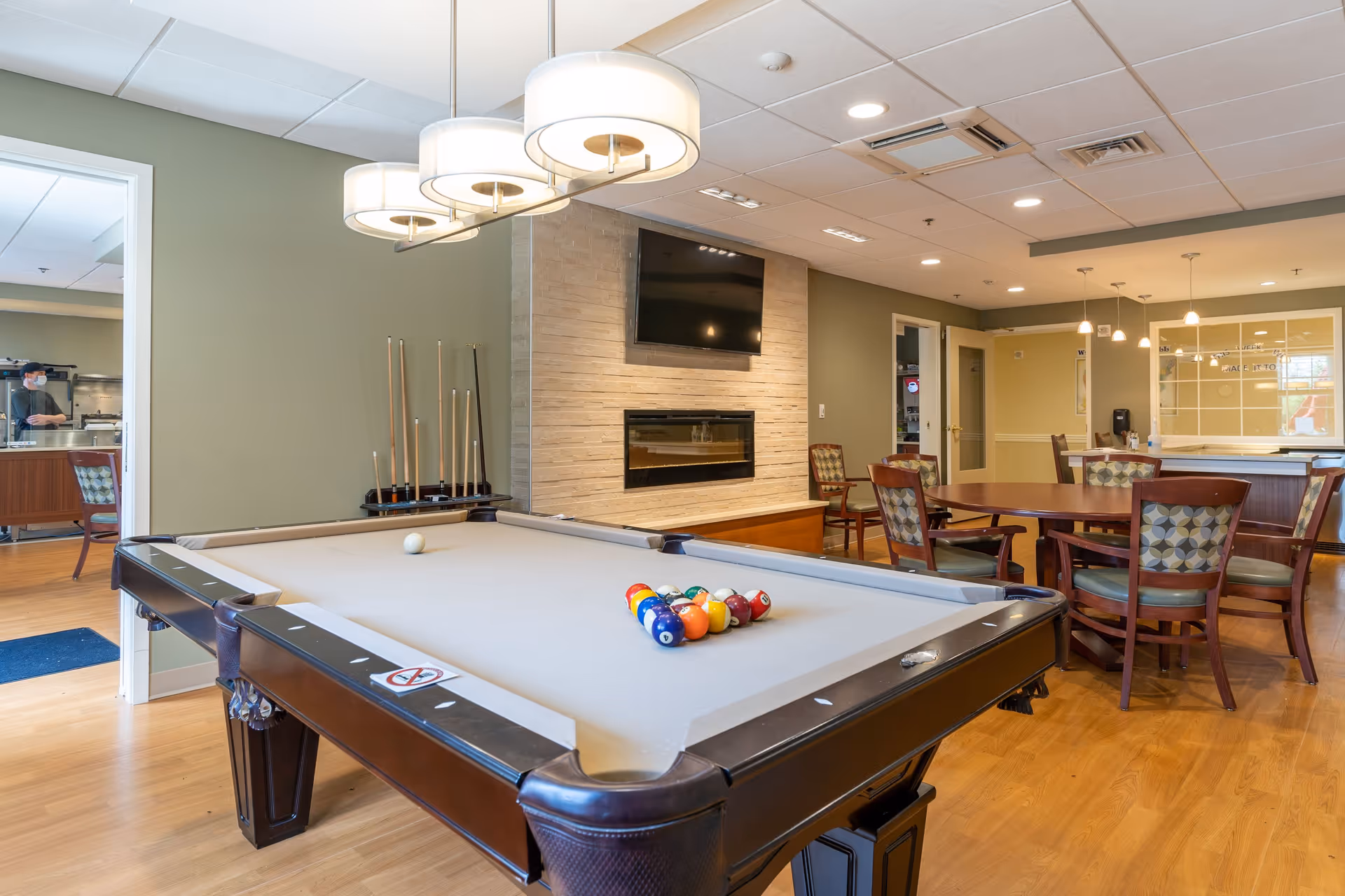 A recreational room at The Park Danforth featuring a pool table with billiard balls arranged for a game. Behind the pool table, there is a modern fireplace with a flat-screen TV mounted above it. To the right, there is a round table with chairs, and pendant lights hanging from the ceiling. The room has wooden flooring and a green accent wall. In the background, a person wearing a mask is visible through an open doorway leading to another room.