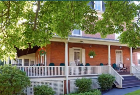 Front exterior view of a brick building with a covered porch featuring several chairs and hanging plants, surrounded by green trees and shrubs.