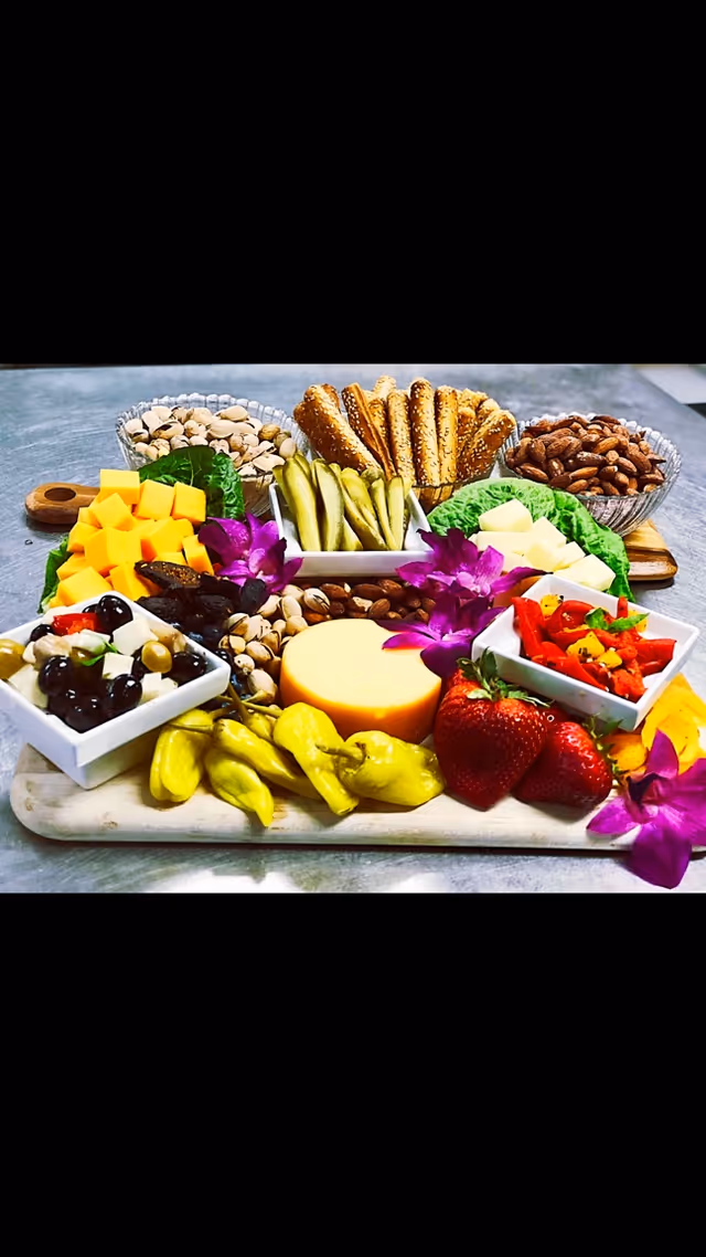 A colorful charcuterie board featuring a variety of foods including cubed cheddar cheese, pickles, pretzel sticks, almonds, pistachios, strawberries, pepperoncini peppers, mixed olives, cubes of white cheese, roasted red peppers, and garnished with purple edible flowers, all arranged on a wooden board with small bowls and lettuce leaves.