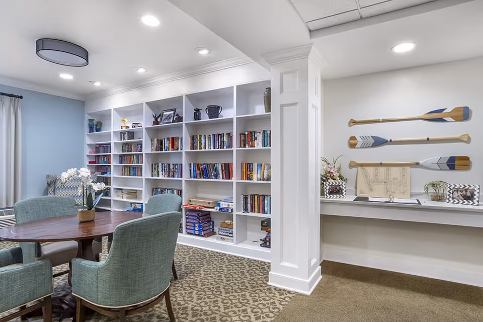 A cozy interior room with a round wooden table surrounded by four upholstered chairs. Behind the table is a large white bookshelf filled with books and board games. To the right, there is a white column and a narrow shelf displaying decorative items including three wooden oars mounted on the wall, framed pictures, and plants. The room has a patterned carpet and soft lighting from ceiling fixtures.