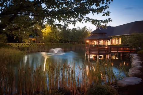 A serene pond at dusk with a water fountain in the center, surrounded by tall grasses and trees. To the right, there is a wooden gazebo-like structure with warm lights, connected to a building partially visible in the background.