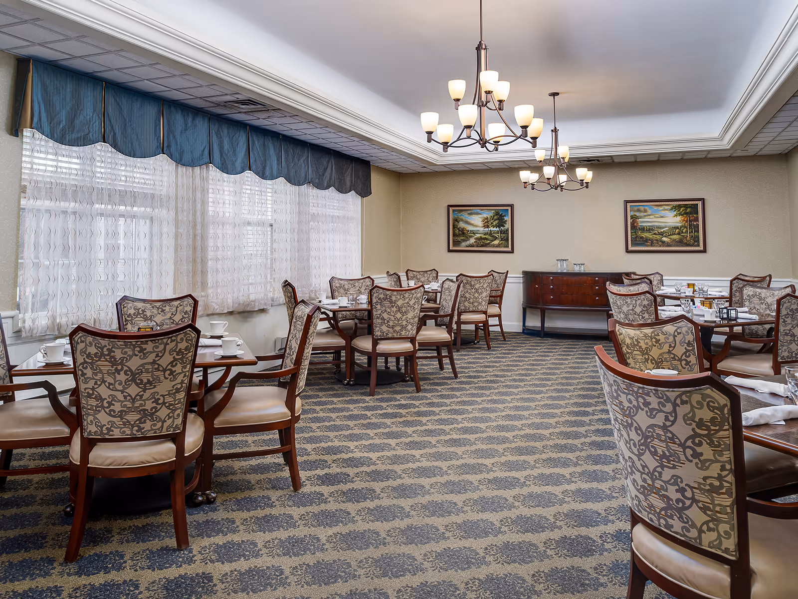 Elegant senior dining room with multiple set tables and upholstered chairs, chandeliers, carpeted floor and large curtained windows.