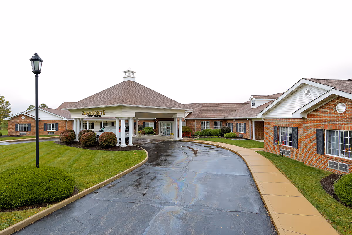 Exterior view of Crown Pointe Assisted Living facility showing a curved driveway leading to a covered entrance with white columns. The building is made of red brick with white trim and has a well-maintained lawn and bushes around it. A black street lamp is visible on the left side of the driveway.