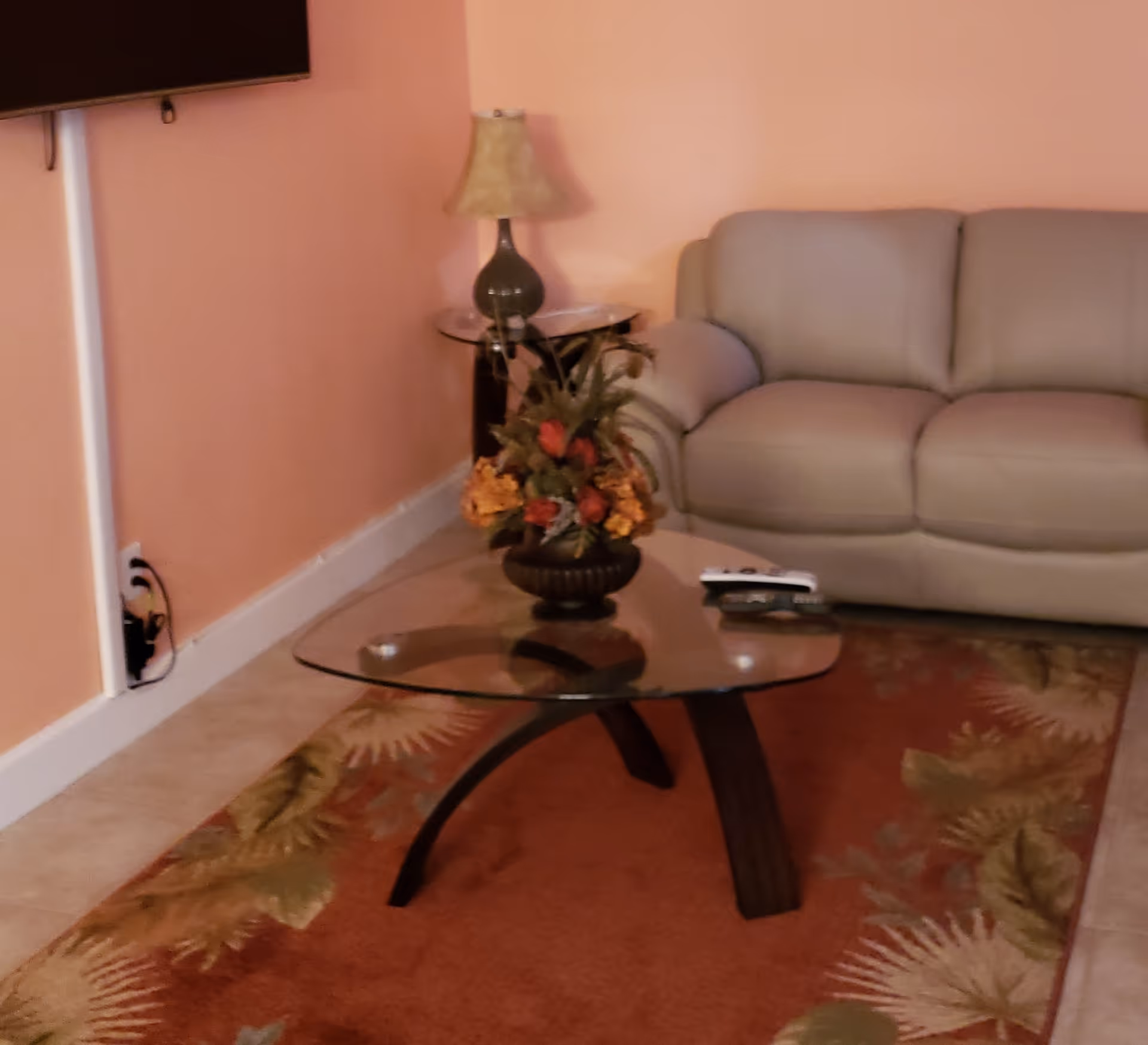 A cozy living room with a beige leather sofa, a glass coffee table with a floral arrangement, a side table with a lamp, and a wall-mounted TV. The room has peach-colored walls and a patterned area rug with leaf designs.