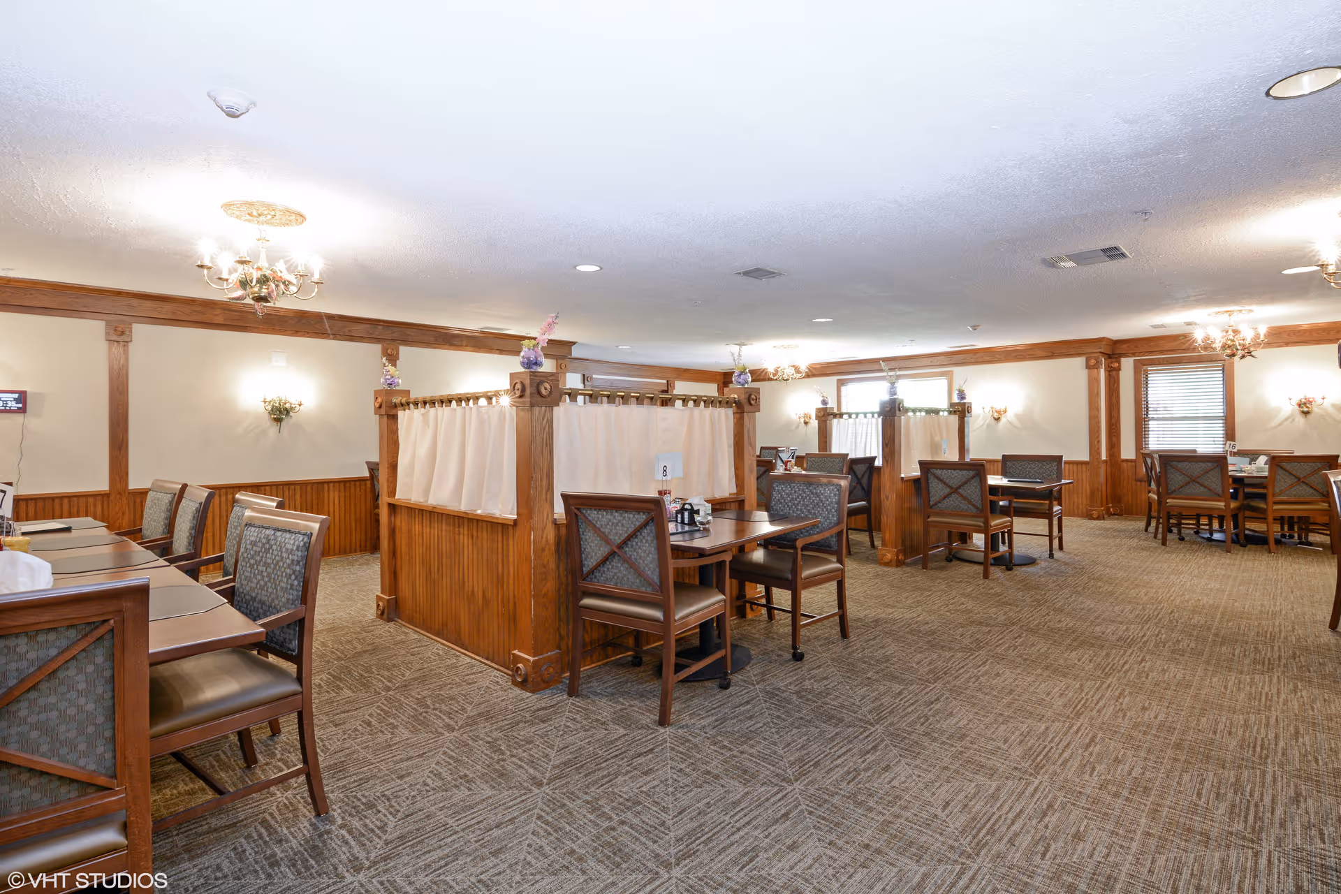 Dining room with wooden partitions, multiple tables and upholstered chairs under chandeliers.