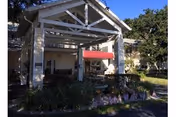 Entrance of a residential building with a covered porch supported by stone pillars, surrounded by greenery and plants under a clear blue sky.