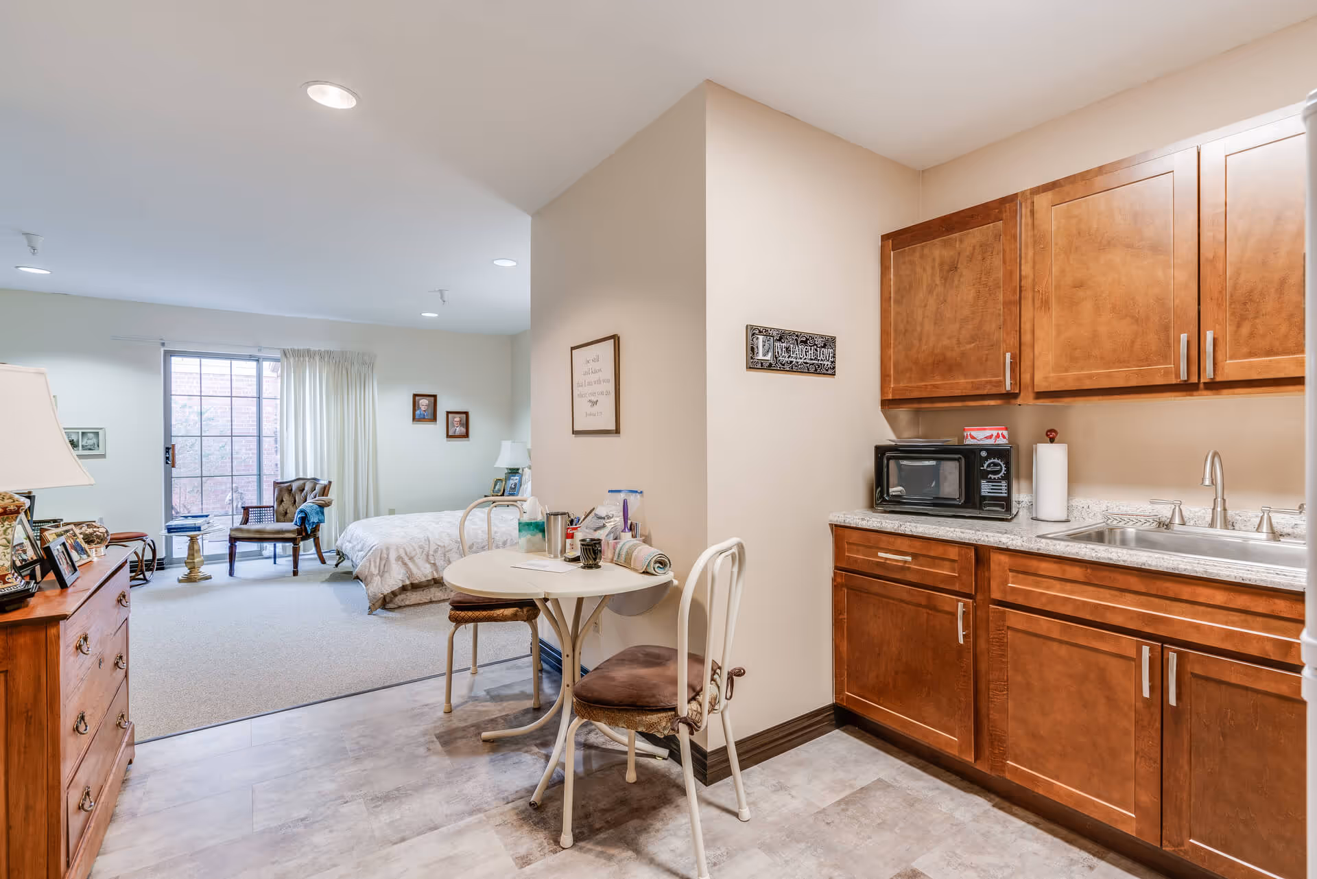 Interior view of a senior living facility room showing a small kitchenette with wooden cabinets, a microwave, and a sink on the right. A small round table with two chairs is positioned near the kitchenette. In the background, there is a bedroom area with a bed, a chair, a dresser with framed photos, and a large window with curtains letting in natural light.