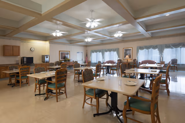 A spacious dining room with multiple tables and chairs arranged neatly. Each table is set with plates, glasses, and utensils. The room has large windows with curtains allowing natural light to fill the space. Ceiling fans with lights hang from the coffered ceiling. There are framed paintings on the walls and a few sofas near the windows.