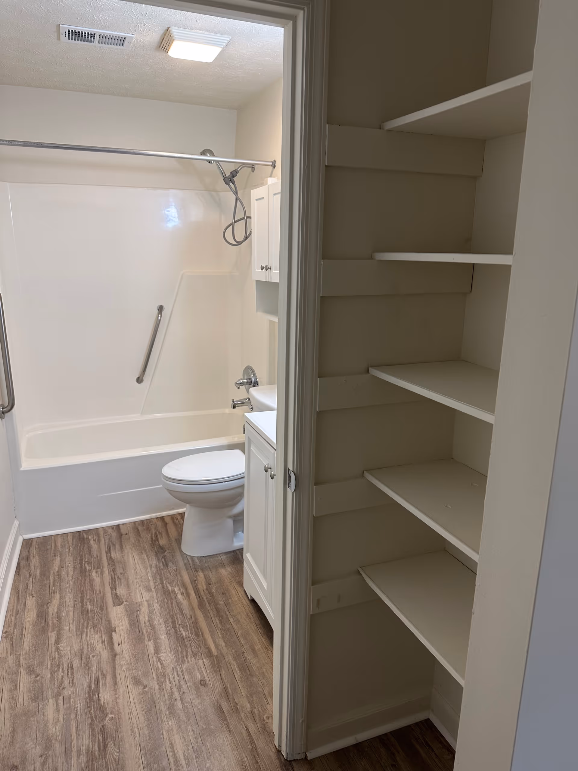 View into a bathroom featuring a white bathtub with a showerhead and grab bar, a white toilet, and a white vanity with a sink. The bathroom has wood-look flooring and a ceiling light. To the right of the doorway, there are built-in white shelves.