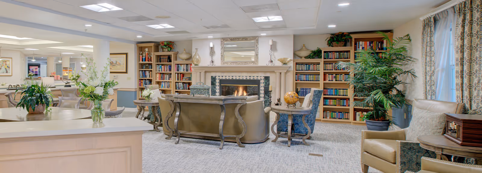 A cozy living room area in a senior living facility featuring a fireplace with a mirror above it, flanked by bookshelves filled with books and decorative items. The room has comfortable seating including a sofa and armchairs, side tables with plants and decorative objects, and large windows with patterned curtains allowing natural light to enter.