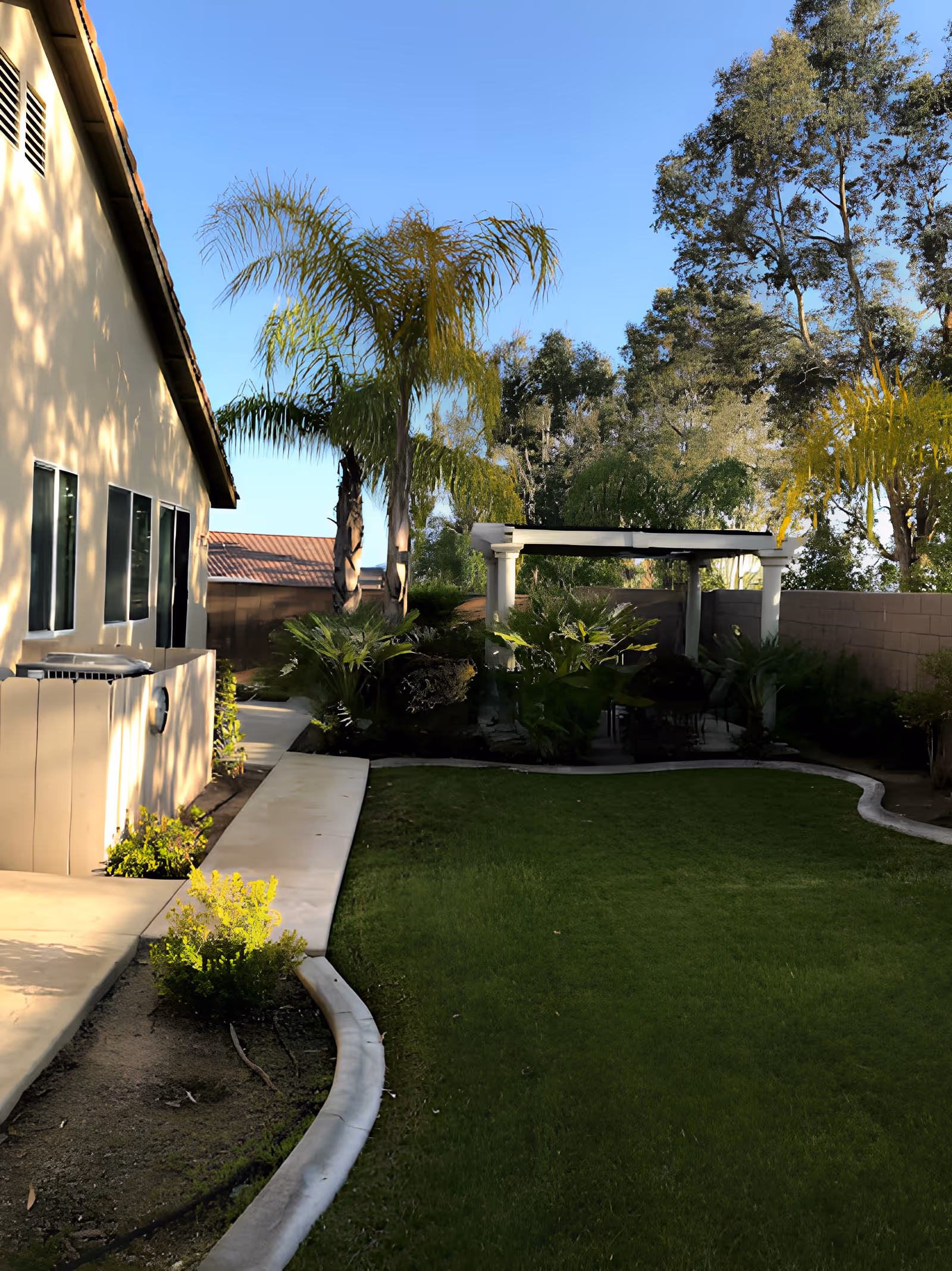 Outdoor garden area with a green lawn, palm trees, and other plants. A beige building with windows is on the left side, and a white pergola with seating is visible in the background. The sky is clear and blue.