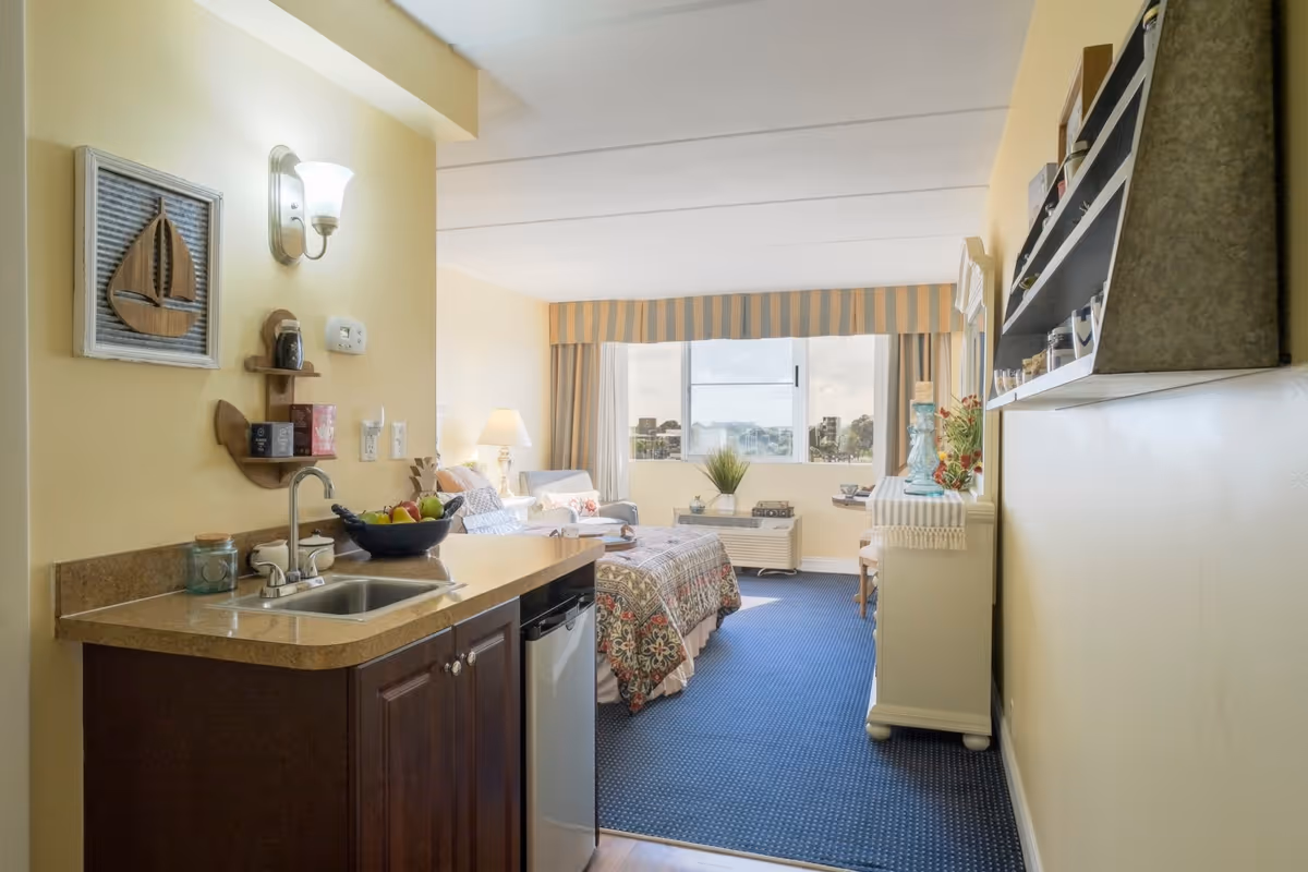 Interior view of a senior living facility room featuring a small kitchenette with a sink and mini fridge on the left, a bed with patterned bedding in the center, a sitting area with a lamp and chair near a large window with striped valance, and a white dresser with decorative items on the right.