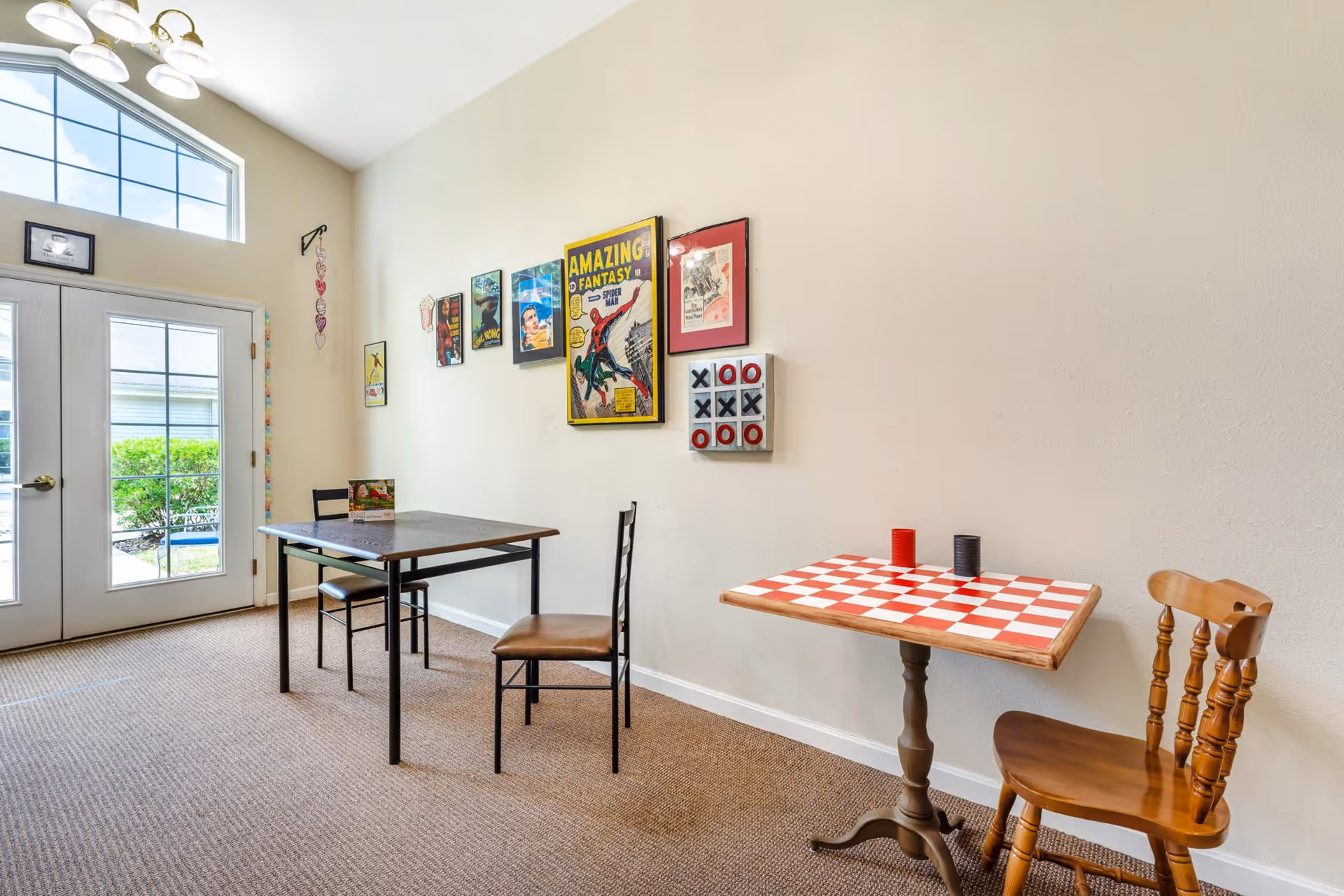 Bright interior common room with two small tables and chairs, wall artwork, and glass double doors to the outside.