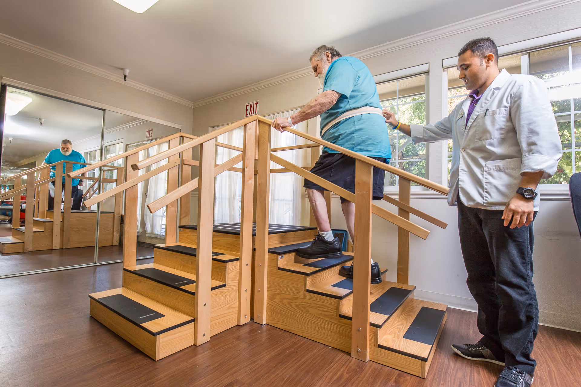 An elderly man wearing a teal shirt and black shorts is walking up a set of wooden stairs with handrails on both sides, assisted by a healthcare professional in a white coat. The room has large windows letting in natural light and a mirrored wall reflecting the scene.