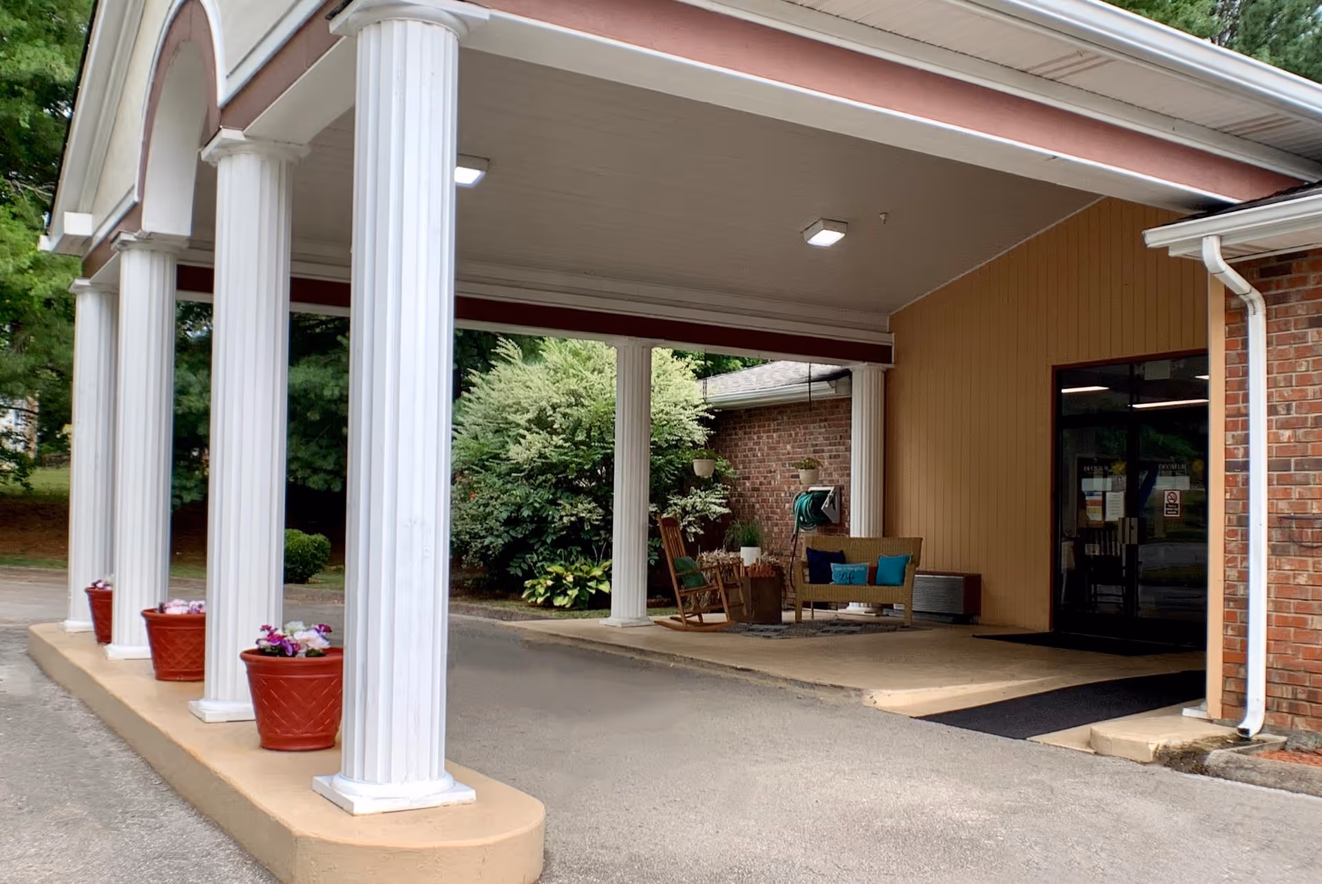 Covered entrance of a brick building with white columns, potted flowers, and outdoor seating by the doorway.