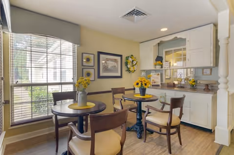 A bright communal dining area with round tables and chairs, sunflower centerpieces, a large window, and a small kitchenette with cabinetry in the background.