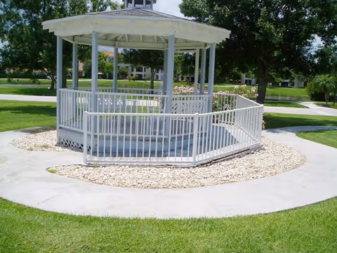 White gazebo with a ramp and railing surrounded by a circular concrete path and green grass in a landscaped outdoor area with trees and buildings in the background.
