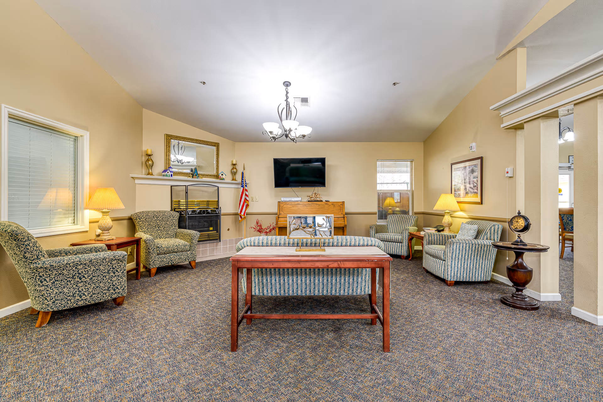 A cozy living room area with patterned armchairs and a striped sofa arranged around a wooden coffee table. The room features a wall-mounted flat-screen TV above a wooden piano, a fireplace with decorative items on the mantel, an American flag, and two windows with blinds. The walls are painted beige, and the floor is carpeted. There are table lamps on side tables providing warm lighting, and a chandelier hanging from the ceiling.