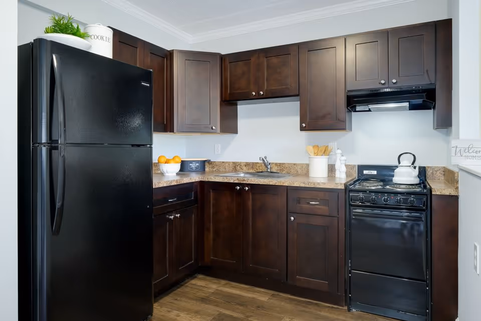 A modern kitchen with dark wooden cabinets, a black refrigerator, a black stove with a white kettle on it, a countertop with a bowl of oranges, a container holding cooking utensils, and a small sink. The floor has a wood-like finish.