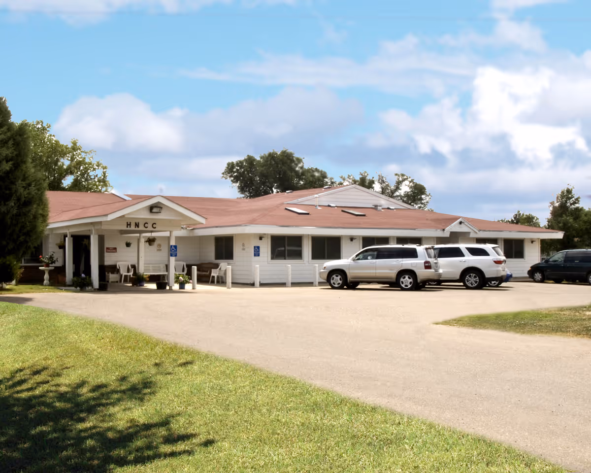 Exterior view of Holly Nursing Care Center, a single-story white building with a red roof and several parked cars in front. The entrance has a covered porch with chairs and potted plants, and the letters HNCC are displayed above the entrance.